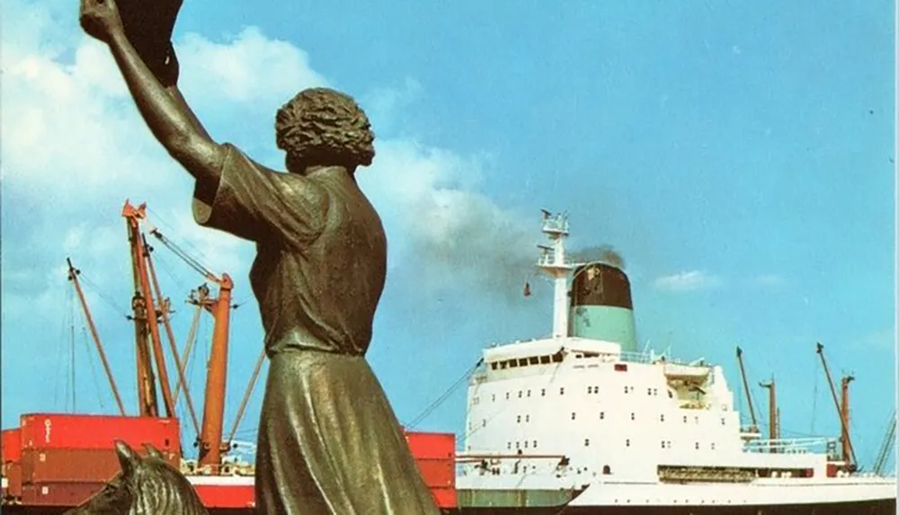 A statue of a person with an upraised arm stands in the foreground overlooking a harbor with a large ship and cranes in the background