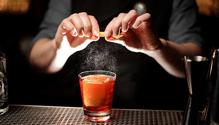 A bartender is deftly twisting an orange peel over a cocktail to spritz its essential oils, enhancing the drink's aroma and flavor.