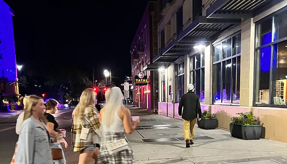 A group of people is walking along a city street at night with colorful lighting from nearby establishments casting a glow on the sidewalk