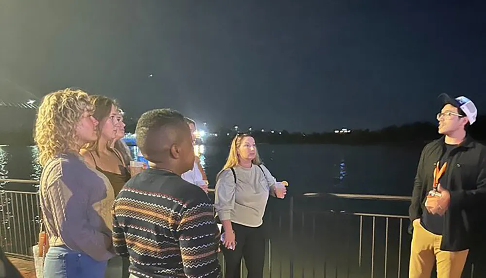 A group of people is gathered near a railing by the water possibly engaged in a conversation during the evening hours