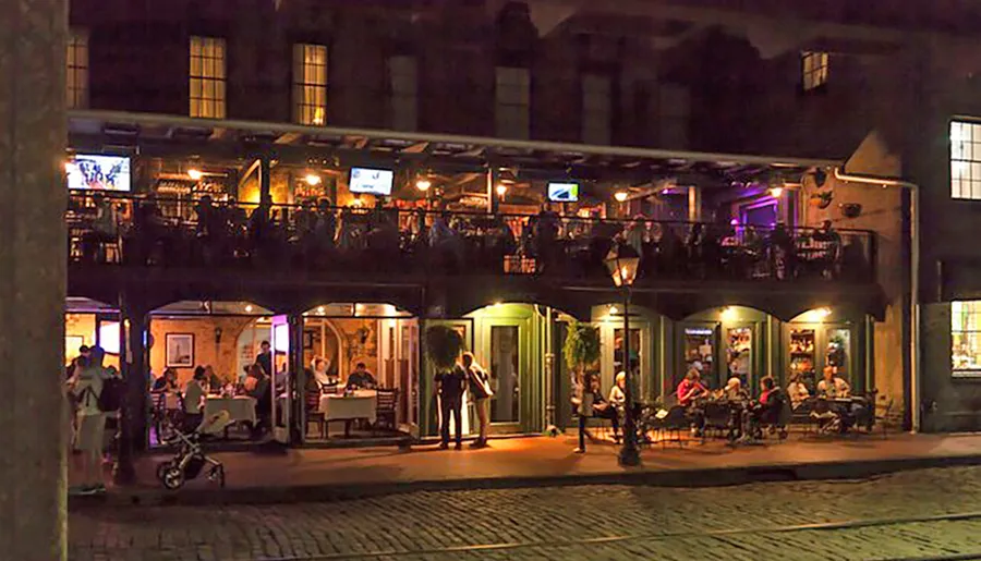 The image portrays a vibrant evening scene at a two-story establishment with patrons dining on the ground level and others enjoying the balcony above, illuminated by warm lights and set against a historic cobblestone street backdrop.