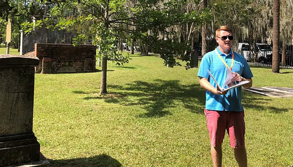 A person is standing in a grassy area near an old structure holding papers and speaking possibly giving a tour or presentation