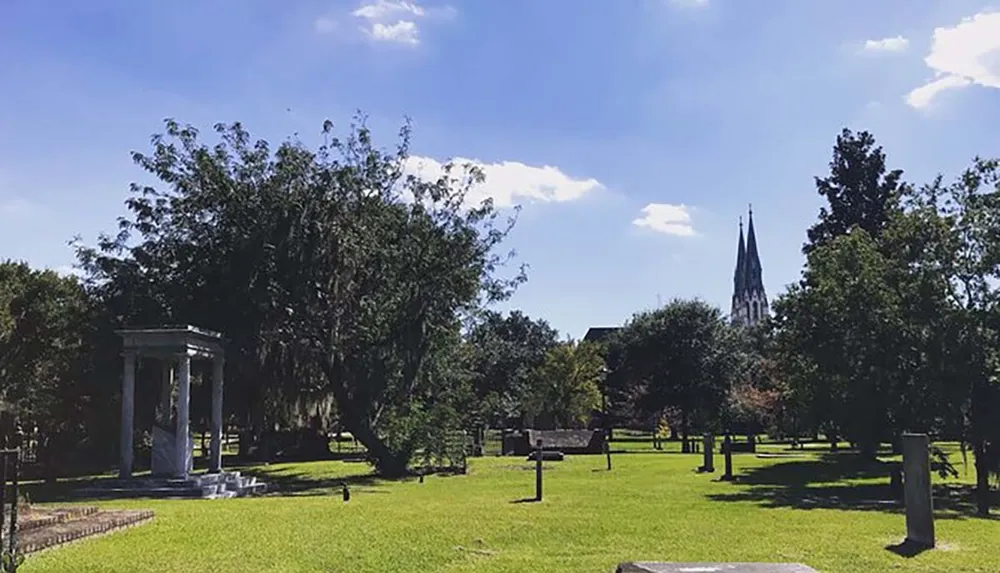 The image depicts a serene park with lush trees a sunlit lawn and a classical column structure with a church spire visible in the background against a clear blue sky