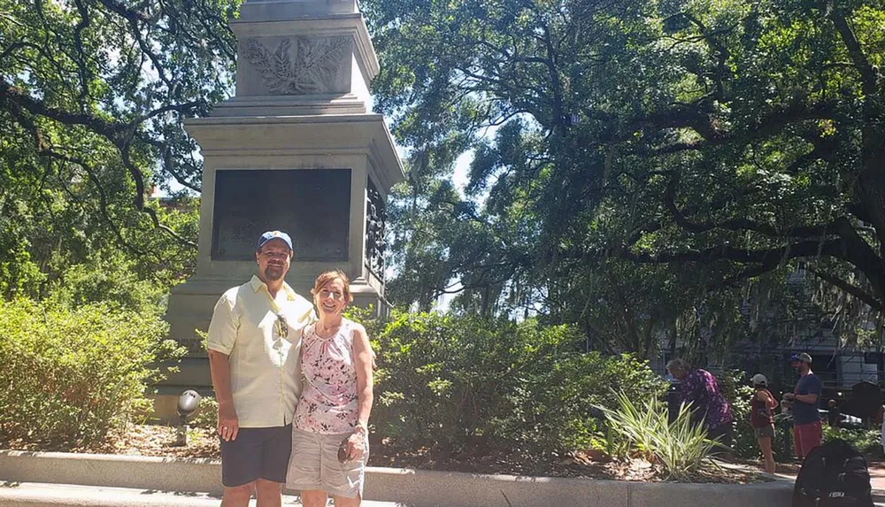 Two people are smiling for a photograph in front of a monument surrounded by lush greenery on a sunny day