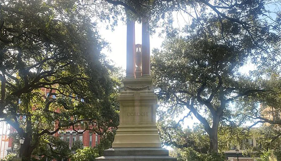 The image features a tall monument surrounded by lush trees with the word GORDON inscribed at the base, set against a backdrop of buildings partially obscured by foliage.