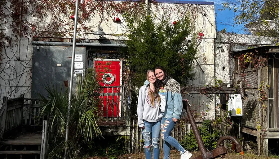 Two smiling individuals are posing in front of a rustic building with a red door and vines, under a clear sky.