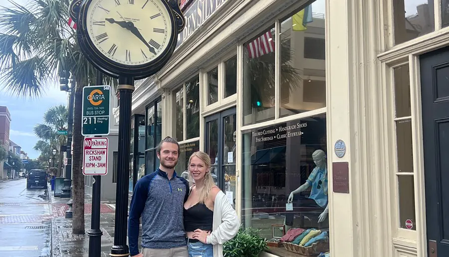 A man and a woman are posing for a photo on a city street with a prominent clock and street signs, outside an elegant clothing store.