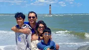 A family of four is smiling for a photo on a sunny day at the beach with a lighthouse visible in the background.
