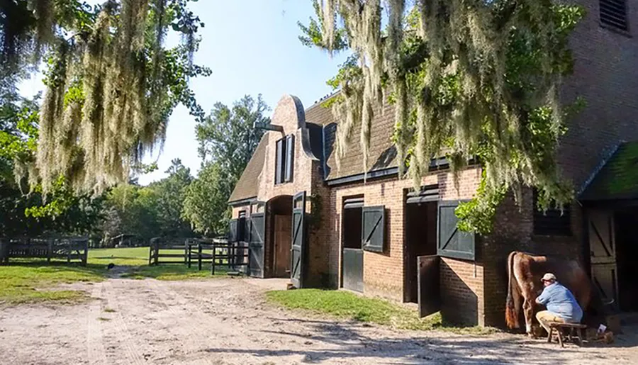 A person sits outside a brick stable under hanging Spanish moss, possibly engaging in a reflective moment or a work-related pause.