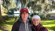Two people are posing for a photo outdoors on a sunny day, with Spanish moss hanging from the trees in the background, suggesting a Southern U.S. setting.