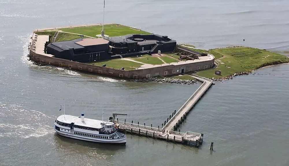 The image is an aerial view of a small fortified island with a pier and a tourist boat docking alongside it