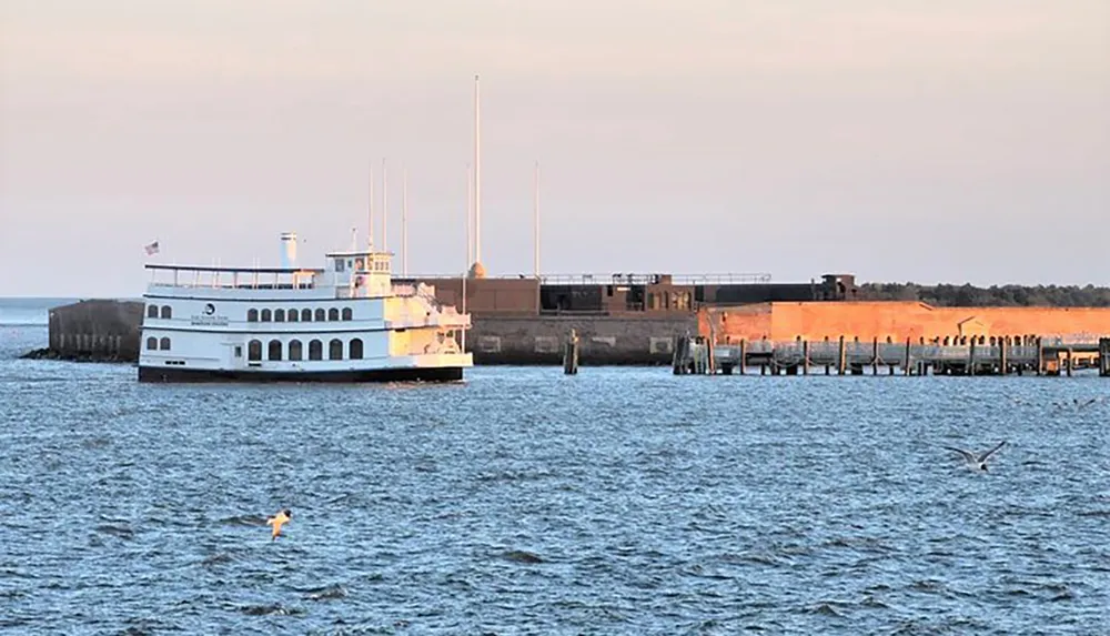 A ferry boat is near a dock by the water at dusk with birds flying nearby and a historical fort structure in the background