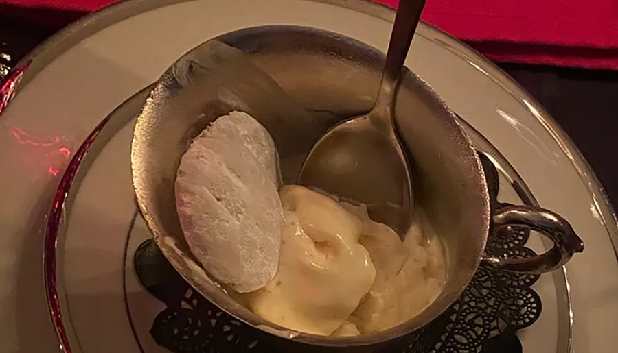 A scoop of ice cream is served beside a warm dessert in a metal bowl with a spoon, set on an elegant table with a red napkin.