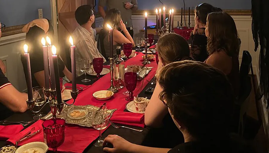 Guests are seated around an elegantly set table with red accents and lit candles, creating an intimate dinner party atmosphere.