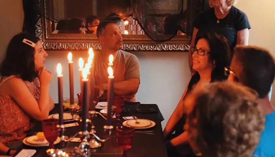 A group of people are engaged in conversation at a dimly-lit dinner table set with lit candles and red glasses, creating an intimate atmosphere.