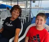 A smiling woman and a young boy are seated on a boat with a view of a large cruise ship in the background