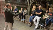 A person is gesturing enthusiastically while speaking to a group of smiling people casually seated on a brick ledge outdoors at night.