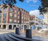 A couple walks past a playful water fountain set against a backdrop of elegant brick buildings under a clear blue sky