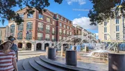 A couple walks past a playful water fountain set against a backdrop of elegant brick buildings under a clear blue sky.
