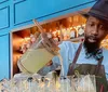 A person wearing a hat and bow tie is skillfully pouring a cocktail into a line of glasses at a bar with a backdrop of neatly arranged bottles