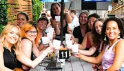 A group of people is smiling at the camera while holding up drinks at an outdoor table marked with the number 11.