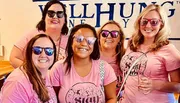 A group of five women wearing pink t-shirts and sunglasses are smiling in front of a sign that says 