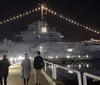 A group of people is strolling along a waterfront promenade at night with a large naval warship adorned with string lights in the background