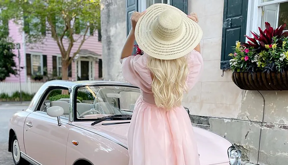 A woman in a pink dress and a straw hat stands facing away from the camera adjusting her hat with a vintage pink car in the foreground and pastel-hued houses in the background