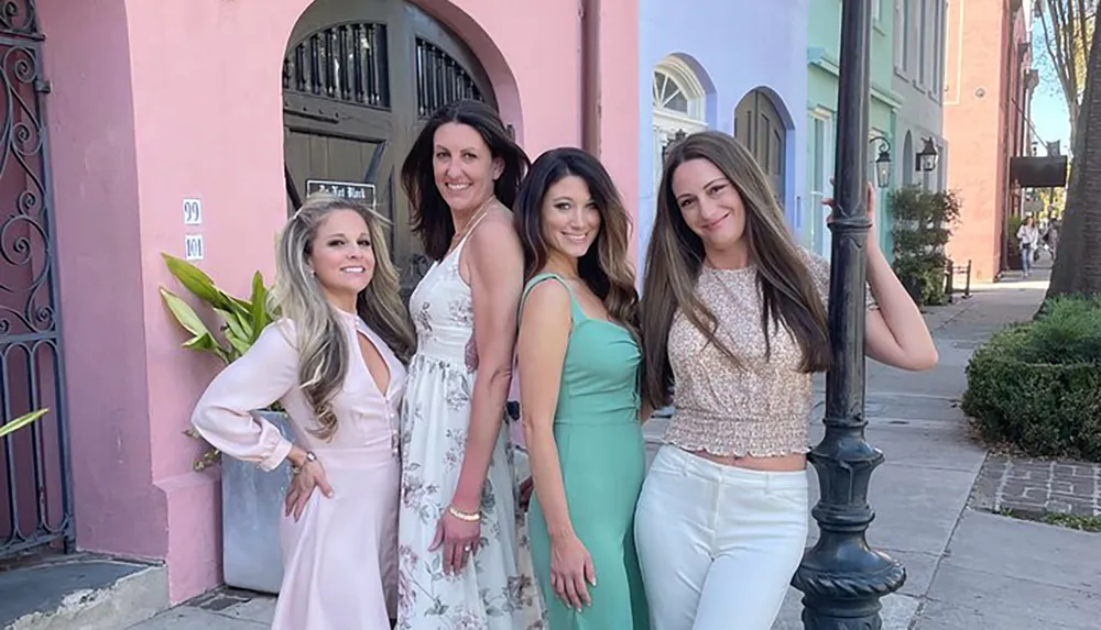 Four women are posing together for a photo on a sunny street smiling and dressed in stylish outfits