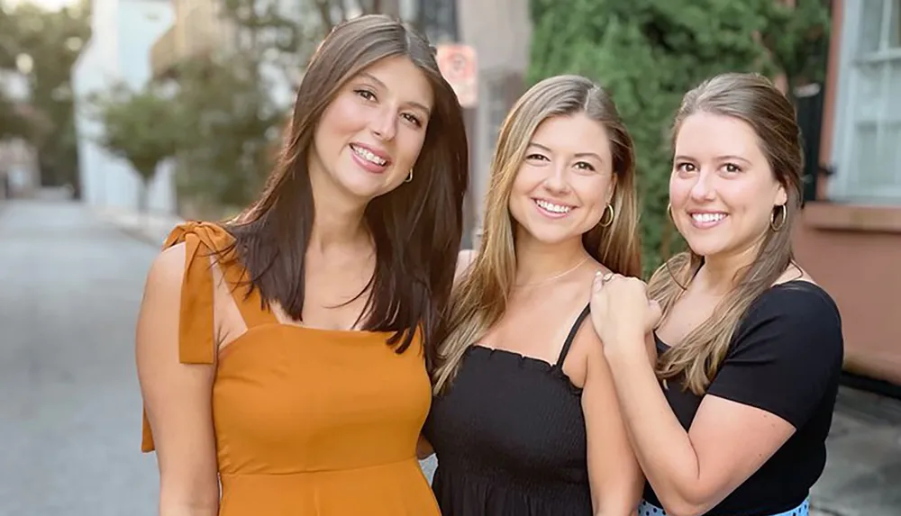 Three smiling women are standing close together on a city street posing for a photograph