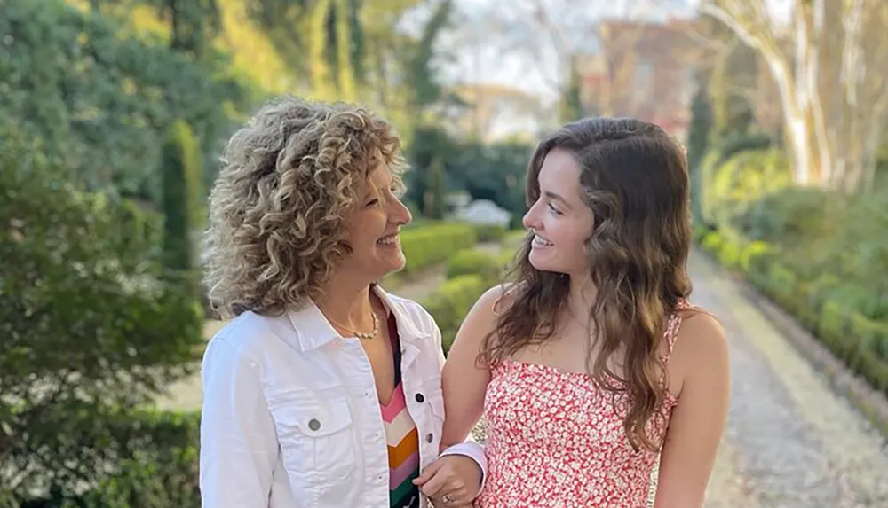 Two happy women are smiling at each other on a sunny park pathway surrounded by greenery