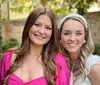 Two smiling women are posing for a photo outdoors with one wearing a pink dress and the other a white dress with a headband both with a backdrop of greenery