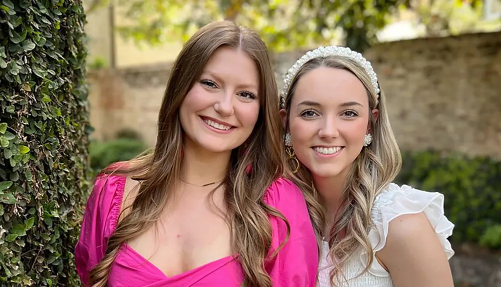 Two smiling women are posing for a photo outdoors with one wearing a pink dress and the other a white dress with a headband both with a backdrop of greenery