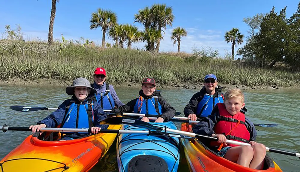 A group of people including children and adults are enjoying a kayaking adventure together in a scenic waterway with lush greenery and palm trees in the background