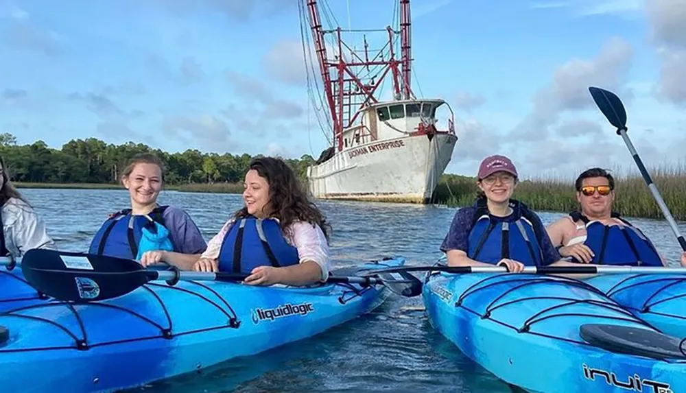 Four people in life vests are smiling and kayaking on a river with a large rusted boat named Judith Enterprise in the background