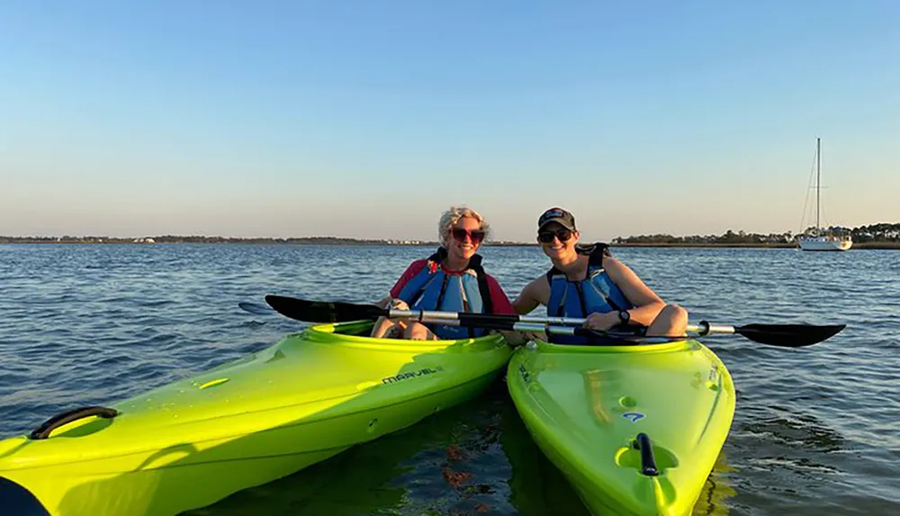 Two people are smiling while kayaking on a calm body of water in the evening light with a sailboat visible in the distance