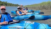 A group of smiling people are enjoying kayaking together on a sunny day in a calm waterway with grassy banks.