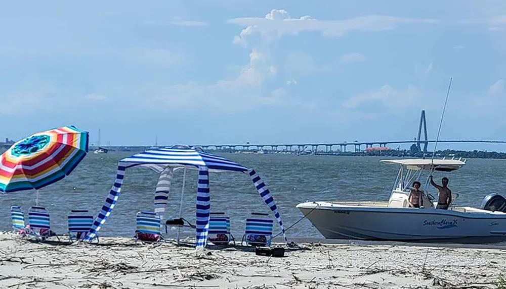 A beach setup with striped chairs and a canopy a colorful umbrella and two individuals on a boat with a bridge in the background