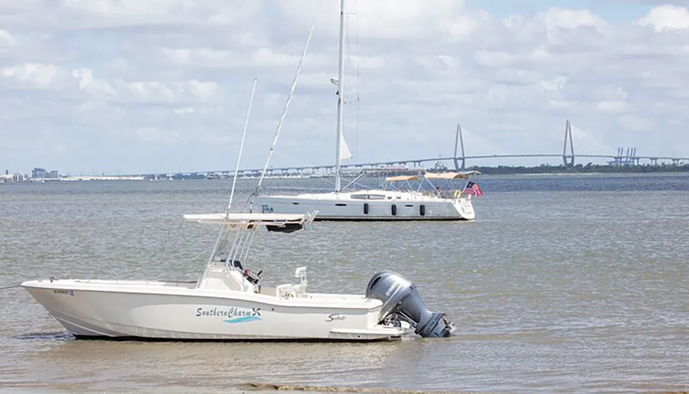 A motorboat is anchored in the foreground on a calm body of water with a sailboat passing by in the background all set against a backdrop of a distant bridge and cloudy sky