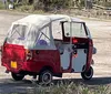 Two individuals are smiling inside a quirky red and white three-wheeled vehicle parked in a driveway with brick detailing in front of a house with a garage door and a landscaped front yard