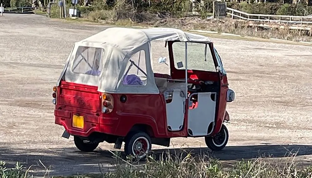 The image shows a small red three-wheeled vehicle with a canvas roof parked on a paved area beside some grass