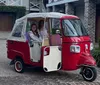 Two individuals are smiling inside a quirky red and white three-wheeled vehicle parked in a driveway with brick detailing in front of a house with a garage door and a landscaped front yard