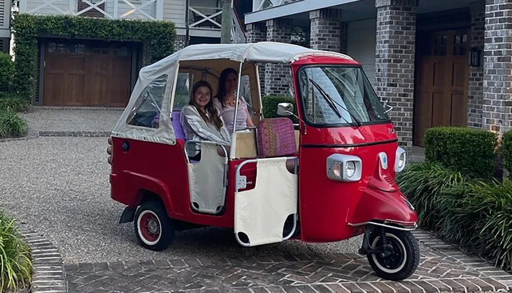 Two individuals are smiling inside a quirky red and white three-wheeled vehicle parked in a driveway with brick detailing in front of a house with a garage door and a landscaped front yard