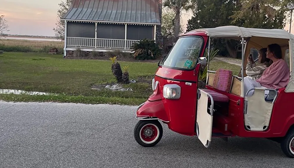 A person is sitting inside a red and white three-wheeled vehicle possibly a tuk-tuk or auto-rickshaw with the door open parked on a road near a body of water and a building with both the driver and a passenger looking at a phone