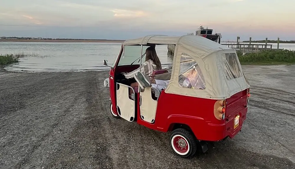 A person sits in a vintage red and white microcar with open doors by a calm waterfront during dusk