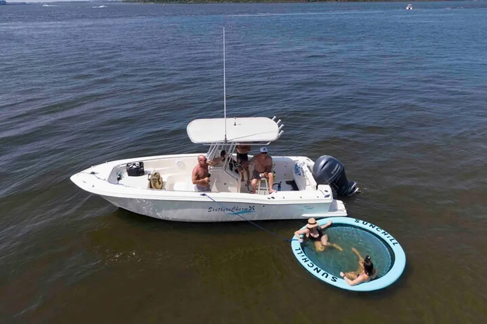 Two people are sitting inside a boat with fishing rods and another person is lounging on a floating ring in the water nearby