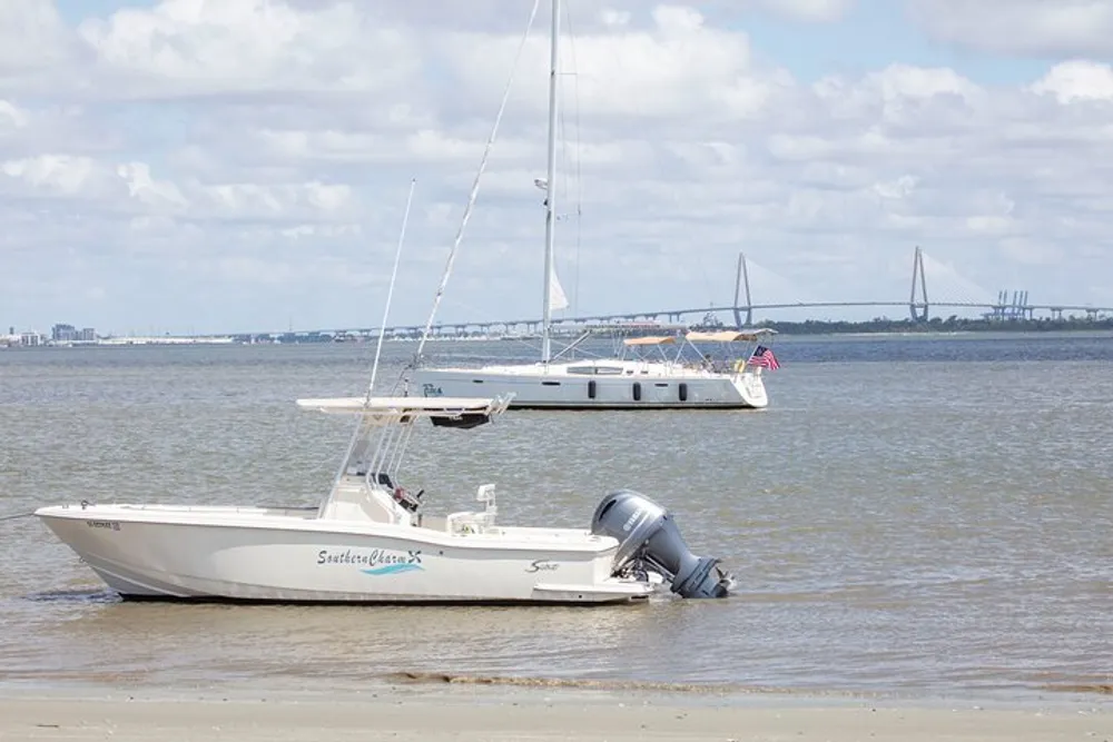 A motorboat is beached on the shore with a sailboat and a bridge in the background under a cloudy sky