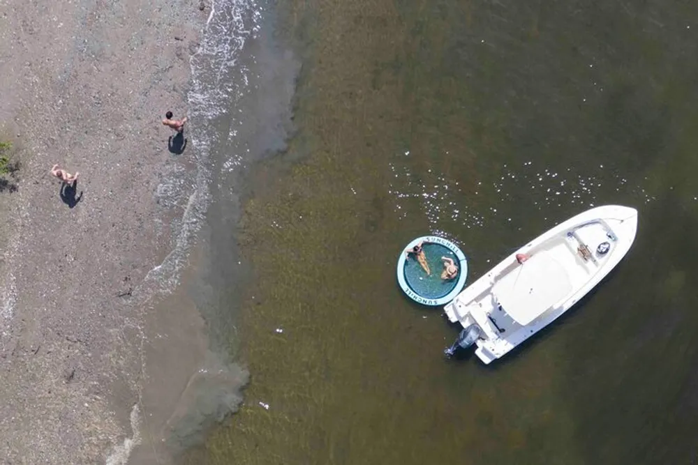 An aerial shot captures two people wading in shallow waters near a docked boat and a person lounging in a small circular floatation device