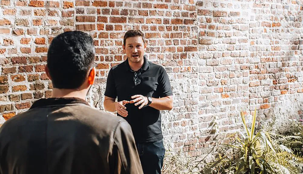 A man gestures while talking to another person in front of a brick wall possibly engaging in a casual outdoor conversation or discussion