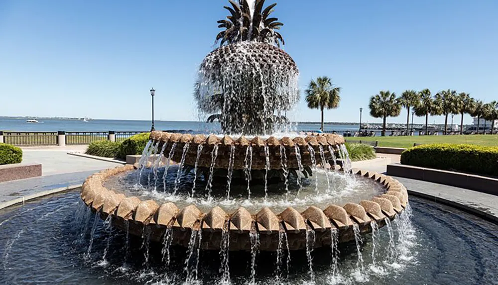 The image shows a multi-tiered pineapple-shaped fountain set in a park with a waterfront promenade and palm trees in the background